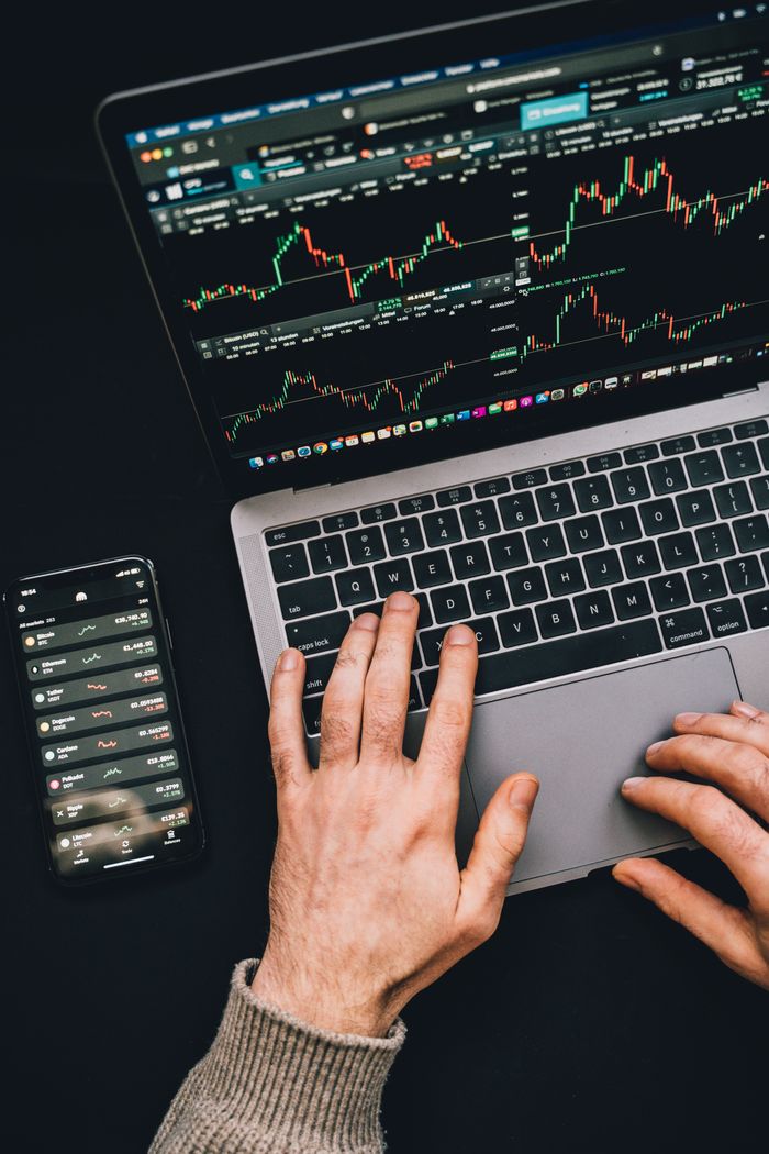 Des mains saisissant un ordinateur portable affichant des graphiques de cryptomonnaies, avec l’écran d’un smartphone visible, dans un contexte axé sur la finance numérique.
